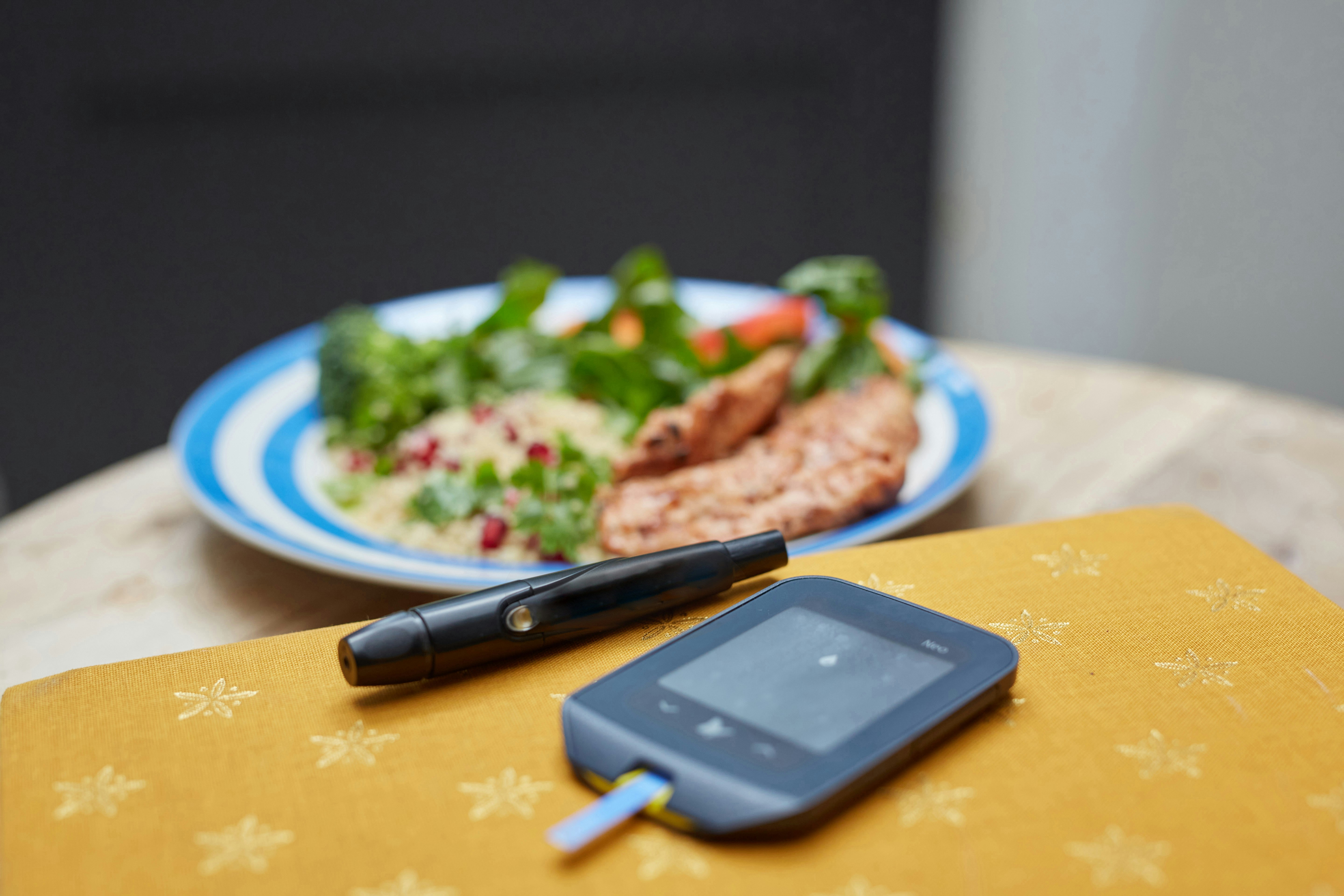 Glucometer and an insulin pen in front with a healthy-looking meal slightly out of focus behind it